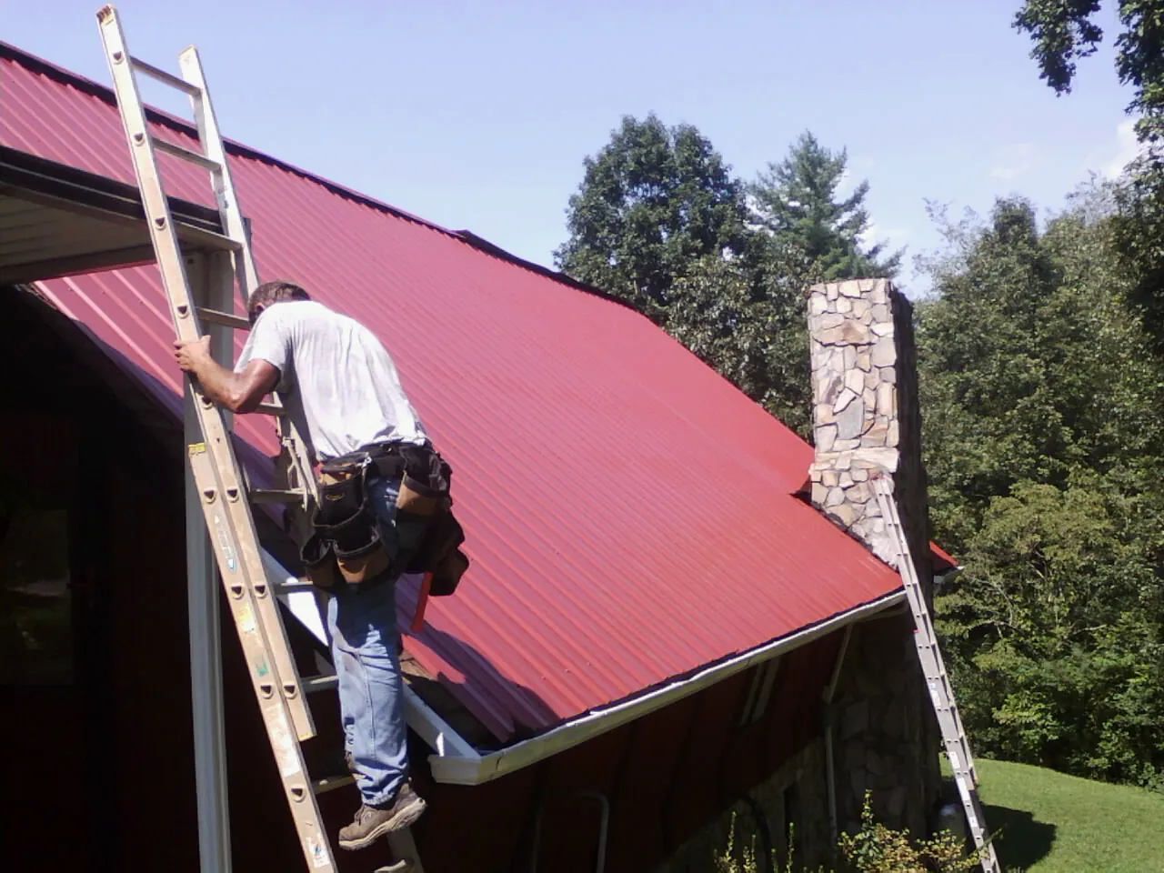 Roofer on a ladder beside a red metal roof and stone chimney — Soffit & Fascia Repair by Roofing Brothers in Cinebar